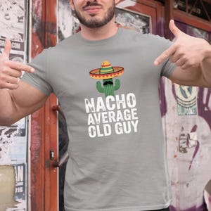 May include: A light gray t-shirt with a graphic of a cactus wearing a sombrero and a mustache. The text "NACHO AVERAGE OLD GUY" is printed below the graphic in white block letters.