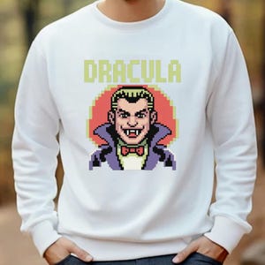 Può includere: Felpa bianca con grafica pixelata di Dracula e la scritta "DRACULA" sopra. La grafica mostra Dracula con colletto viola, papillon rosso e sfondo rosso, in stile videogioco retrò.