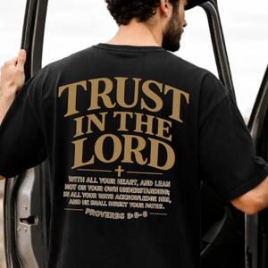 May include: Black t-shirt featuring the gold text "TRUST IN THE LORD" and a quote from Proverbs 3:5-6. The shirt has a casual style and is suitable for everyday wear.