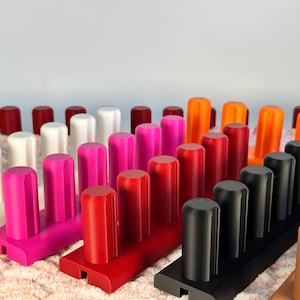 May include: A collection of colorful knitting looms in various shades, including white, pink, red, black, and orange. Each loom has a series of pegs for crafting. The looms are arranged on a textured, light-colored surface.