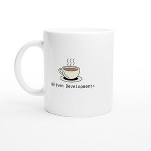 Peut inclure: Une tasse à café en céramique blanche avec une illustration de dessin animé d'une tasse de café fumante et le texte "<Driven Development>" imprimé sur le devant.