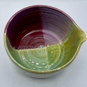May include: A handmade ceramic mixing bowl with a pouring spout. The bowl is divided into two color sections: a deep burgundy and a light green. The rim has a purple hue. The bowl is ideal for kitchen use.