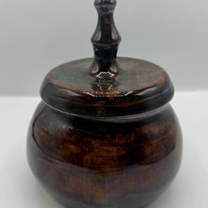 Könnte beinhalten: Ein kleines, rundes, braunes Keramikgefäß mit Deckel. Das Gefäß hat eine glänzende Oberfläche und eine dunkelbraune Farbe mit hellbraunen Streifen. Der Deckel hat einen Knopf oben. Das Gefäß wird wahrscheinlich zur Aufbewahrung oder Dekoration verwendet.