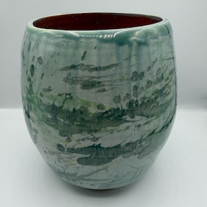 May include: A ceramic vase with a rounded shape and a wide opening. The exterior is a light blue-green with splattered green and gray accents. The interior is a deep reddish-brown color.