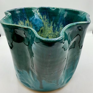 May include: A handcrafted ceramic vase with a wavy rim. The vase has a glossy, teal glaze with streaks of blue and yellow, creating a unique, artistic appearance. The vase is a decorative item.