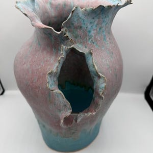 May include: A handcrafted ceramic vase with a unique, organic shape. The vase features a blend of pink and teal glazes, with a textured surface and an irregular opening. The rim has a ruffled edge, adding to its artistic appeal.