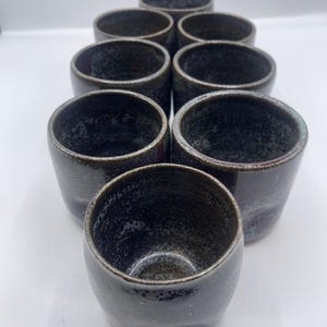 Könnte beinhalten: Ein Set aus acht dunkelgrauen Keramikbechern. Die Becher haben eine gesprenkelte Textur und eine leicht unregelmäßige Form. Die Ränder sind hellgrau. Die Becher sind in einem versetzten Muster angeordnet, wobei die Öffnungen zum Betrachter zeigen.