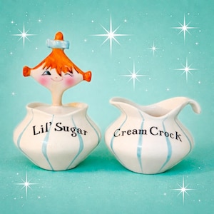 May include: A vintage-style ceramic sugar bowl and creamer set. The sugar bowl is topped with a whimsical face with orange hair and a blue hat. Both pieces are white with blue vertical stripes and the words "Lil' Sugar" and "Cream Crock".