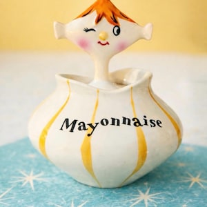 Puede incluir: Un tarro de mayonesa de cerámica de estilo vintage con una cara caprichosa en la parte superior. El tarro es blanco con rayas amarillas y la palabra "Mayonnaise" en negro. La cara tiene pelo naranja y guiña un ojo. Sobre una superficie azul.