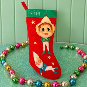 May include: A red felt Christmas stocking featuring a green cuff with the name KIM embroidered in white. The stocking is decorated with a three-dimensional elf face, a flat elf figure, and several small gold star appliques on the red surface.