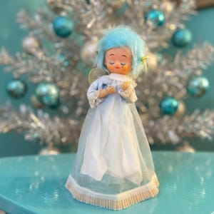 Könnte beinhalten: Vintage Weihnachtsbaum-Engelsfigur mit blauer Perücke und hellblau-weißem Kleid. Der Engel hat geschlossene Augen und kleine goldene Flügel. Das Kleid hat eine plissierte Bordüre. Der Hintergrund zeigt einen silbernen Weihnachtsbaum mit blauen Kugeln.