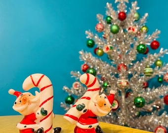 Może przedstawiać: Dwie ceramiczne figurki bożonarodzeniowe w kształcie Świętego Mikołaja i lasek cukrowych. Figurki Świętego Mikołaja noszą czerwone stroje z czarnymi butami i czapkami. W tle srebrna choinka z kolorowymi ozdobami.
