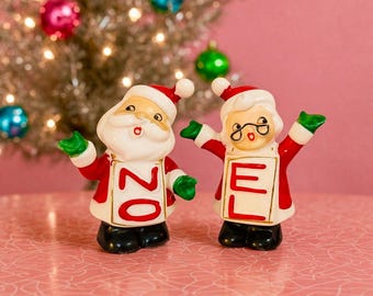 Może przedstawiać: Dwie ceramiczne figurki bożonarodzeniowe, Święty Mikołaj i Pani Mikołajowa, z wyciągniętymi ramionami. Święty Mikołaj trzyma znak z numerem "20", a Pani Mikołajowa trzyma znak z literą "E". Oboje noszą czerwono-białe stroje z zielonymi rękawicami.