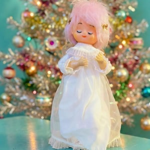 Könnte beinhalten: Ein Vintage-Weihnachtsbaumaufsatz mit einer Puppe mit rosa Haaren und einem weißen Kleid. Die Puppe hat geschlossene Augen und kleine goldene Akzente im Haar. Der Hintergrund zeigt einen silbernen Weihnachtsbaum mit bunten Ornamenten.
