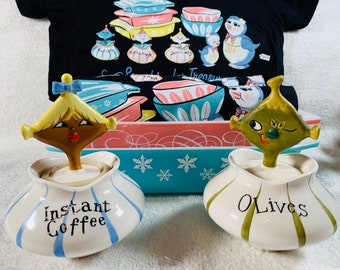 May include: Two ceramic kitchen canisters with whimsical character toppers. The canister on the left is blue and white with the text "Instant Coffee". The canister on the right is green and white with the text "Olives".
