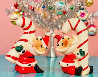 May include: Two ceramic Santa Claus figurines, each wearing a red suit and holding a large, curved candy cane with red and white stripes. The figures have painted facial features, green mittens, and black boots, standing in a pair.