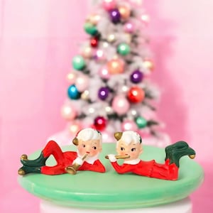 Puede incluir: Dos figuras de duendes navideños de cerámica con trajes rojos y verdes, tumbadas sobre una superficie verde. Un duende toca la trompeta, el otro la flauta. Un árbol de Navidad decorado con adornos coloridos está al fondo.