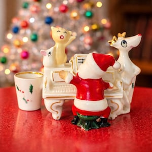 Puede incluir: Un conjunto de figuras navideñas festivas con Papá Noel tocando el piano, un gato cantando y un reno. Las figuras son blancas con detalles en rojo y dorado. También hay una pequeña taza con acebo y bayas.