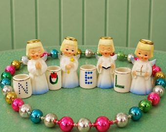 May include: A set of four ceramic angel figurines with blonde hair and white robes, each paired with a small cylindrical candle holder. The holders feature the letters N, O, E, L in blue and green. A string of colorful glass beads encircles them.