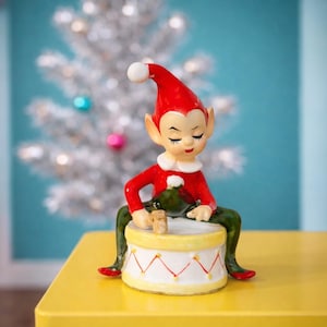 Puede incluir: Una figura de elfo navideño vintage. El elfo lleva un atuendo rojo con cuello blanco y un gorro rojo a juego con un pompón blanco. El elfo está sentado en un tambor amarillo y blanco, con pantalones y zapatos verdes. El fondo incluye un árbol de Navidad plateado.