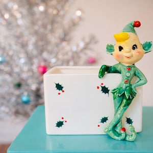 Könnte beinhalten: Eine grüne Keramik-Weihnachtself-Figur mit gelber Mütze und roten Akzenten, die sich an einen weißen Pflanztopf lehnt. Der Pflanztopf ist mit Stechpalmen und roten Beeren verziert. Im Hintergrund befindet sich ein silberner Lametta-Weihnachtsbaum.