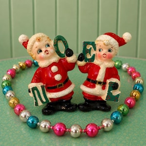 May include: Two vintage ceramic figurines of children dressed as Santa Claus, each wearing a red suit with white trim and a matching hat. They hold green letters spelling NOEL, surrounded by a circular string of colourful metallic beads.