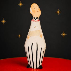 May include: A tall, slender figurine of a bald person with a pale complexion, wearing a white jacket with black vertical stripes at the base, a red and white striped collar, a black bow tie, and holding a blank rectangular beige card.