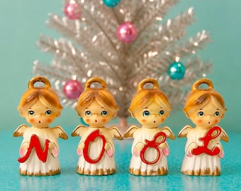 Peut inclure: Quatre figurines d'anges en céramique disposées en rangée, portant chacune une robe blanche avec des bordures dorées et tenant une lettre rouge en cursive. Ensemble, les anges forment le mot NOEL. Chaque ange a des cheveux blonds et des ailes.