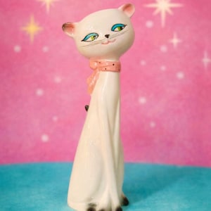 Może przedstawiać: Biała ceramiczna figurka kota z różową chustą i czarnymi akcentami. Kot ma niebieskie oczy, różowy nos i czarne wąsy. Figurka jest w pozycji siedzącej i ma estetykę w stylu mid-century modern.