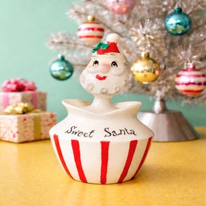 May include: A vintage ceramic Santa Claus figurine with a red and white striped base and the words "Sweet Santa" in black script. The Santa has a red hat and a smiling face. Christmas ornaments and wrapped gifts are in the background.