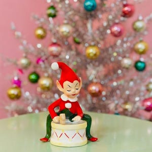 Puede incluir: Una figura de elfo navideño de cerámica con gorro y chaqueta rojos, pantalones verdes y cuello blanco. El elfo está sentado, tocando un tambor con base crema y detalles rojos. Un árbol de Navidad plateado con adornos coloridos en el fondo.