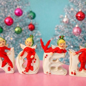 May include: Christmas-themed figurines spelling out the word "NOEL". Each figurine features a blonde elf in a red outfit with white wings, posed on a white base. The background includes silver Christmas trees and colorful ornaments.