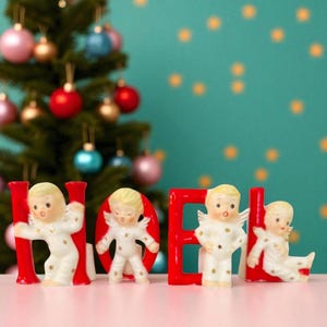 Könnte beinhalten: Weihnachtsdekoration mit den roten Buchstaben "NOEL" und Keramik-Engelsfiguren. Die Engel tragen weiße Pyjamas mit goldenen Sternen. Ein dekorierter Weihnachtsbaum befindet sich im Hintergrund.