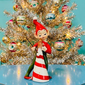 Puede incluir: Una figura de elfo navideño vintage con gorro y chaqueta rojos, pantalones verdes y un cono a rayas blancas y rojas. El elfo sostiene una varita negra. Un árbol de Navidad de oropel plateado con adornos coloridos en el fondo.