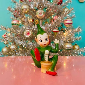 Puede incluir: Figura de elfo navideño de cerámica vintage con atuendo verde, detalles rojos y expresión de sorpresa. El elfo sostiene una vela y está frente a un árbol de Navidad de oropel plateado con adornos coloridos.