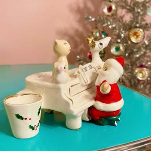 May include: A ceramic white grand piano figurine featuring a seated Santa Claus in a red suit, a small white cat, and a larger cream-colored cat. A separate small white cup with a green holly and red berry pattern sits beside the piano.