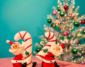 May include: Two ceramic Santa Claus figurines, each wearing a red suit with white trim and black boots, are depicted leaning against large, curved candy canes with red and white stripes. The figures have painted facial features and green mittens.