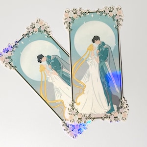 May include: Two rectangular stickers featuring a romantic illustration. The artwork depicts a couple embracing in front of a full moon. The stickers are framed with floral and foliage designs, with a holographic effect on the edges.