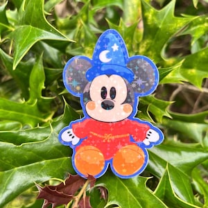 May include: A holographic sticker of Mickey Mouse dressed as a sorcerer. Mickey is wearing a blue wizard hat with a crescent moon and stars, a red robe, and orange shorts. The sticker has a glittery, iridescent effect.