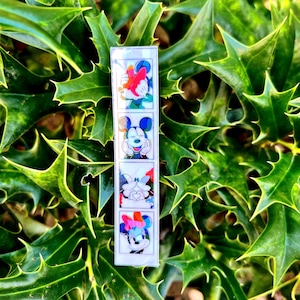 May include: A photo booth strip with four images of Mickey Mouse. The images show Mickey Mouse making different expressions, including a kiss, a wink, and a smile. The strip is white with a black border.