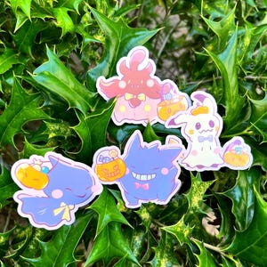 以下が含まれることがあります： ハロウィーンの衣装を着た漫画のキャラクターが描かれた、カラフルなステッカーのコレクション。紫色のゲンガー、カボチャを持ったピンク色のキノコのようなキャラクター、青い鳥のようなキャラクター、白い幽霊のようなキャラクターなどがあります。各キャラクターはバスケットを持っています。