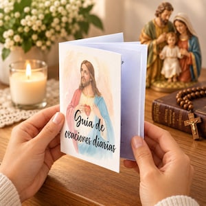 May include: A white prayer booklet titled Guia de oraciones diarias featuring a watercolor illustration of Jesus Christ with a sacred heart. The booklet is held open, displaying a soft blue interior page, with a religious theme and imagery.