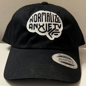May include: Black baseball cap with a white embroidered brain graphic that says "Normalize Anxiety". The cap is adjustable and has a label that says "The Authentic Yupoong Adjustable Yupoong Since 1974".