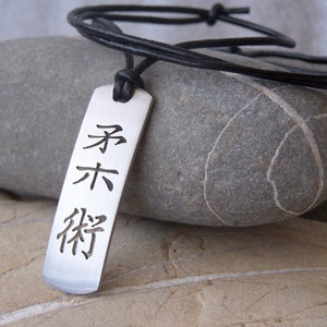 Puede incluir: Un collar con un colgante de plata y un cordón de cuero negro. El colgante es rectangular y presenta tres caracteres chinos:  "宗" "末" "将".