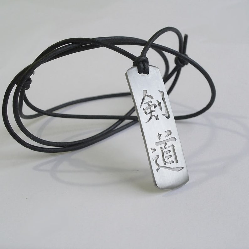 May include: A silver pendant necklace with a black leather cord. The pendant is rectangular and features the Japanese characters "剣道" (kendo) which means "the way of the sword".