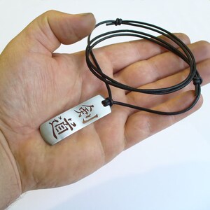 May include: A silver pendant necklace with a black leather cord. The pendant is rectangular and features the Japanese word "道" (pronounced "michi") which means "way" or "path".