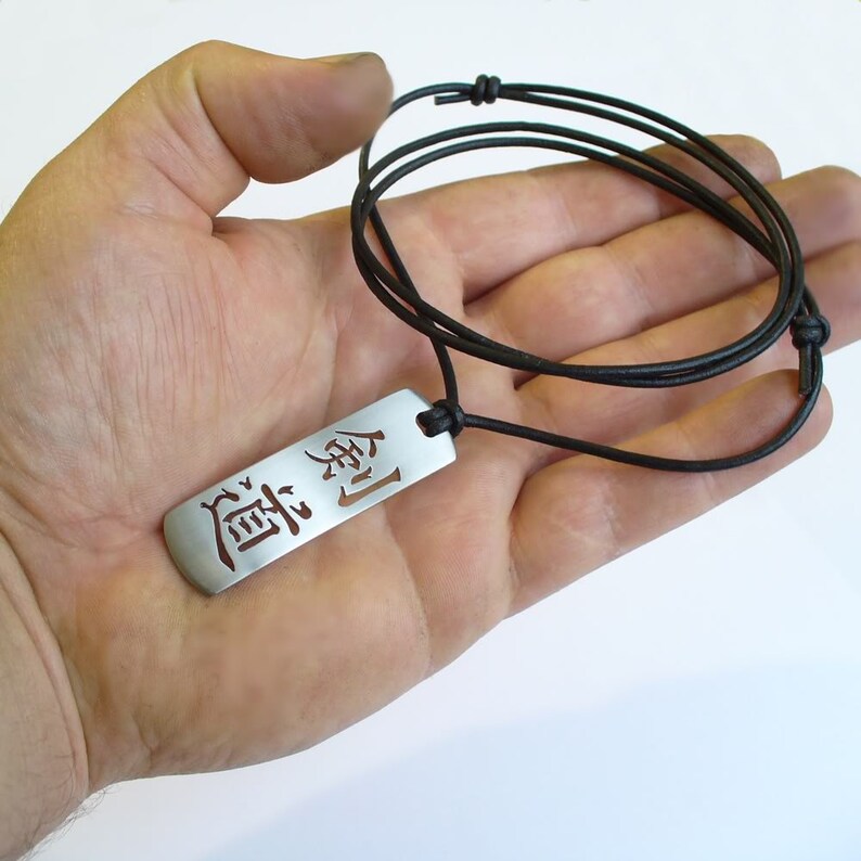 May include: A silver pendant necklace with a black leather cord. The pendant is rectangular and features the Japanese word "道" (pronounced "michi") which means "way" or "path".
