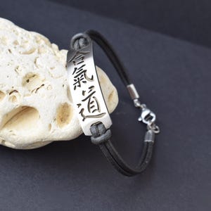 Op de afbeelding: Een zilverkleurige armband met de Japanse Aikido-tekens gegraveerd op een rechthoekige plaat. De armband heeft een donkergrijze leren koord en een zilveren sluiting. Het ontwerp is eenvoudig en elegant.