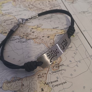 May include: A black leather bracelet with a silver rectangular charm featuring a stylized Japanese kanji character. The bracelet is lying on a map.