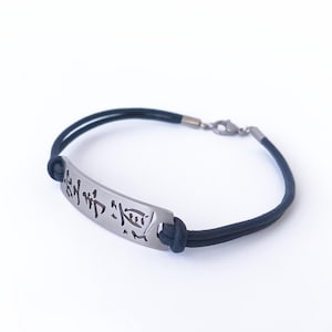 May include: A black cord bracelet with a silver-colored rectangular plate. The plate has etched Japanese characters. The bracelet has a clasp closure.